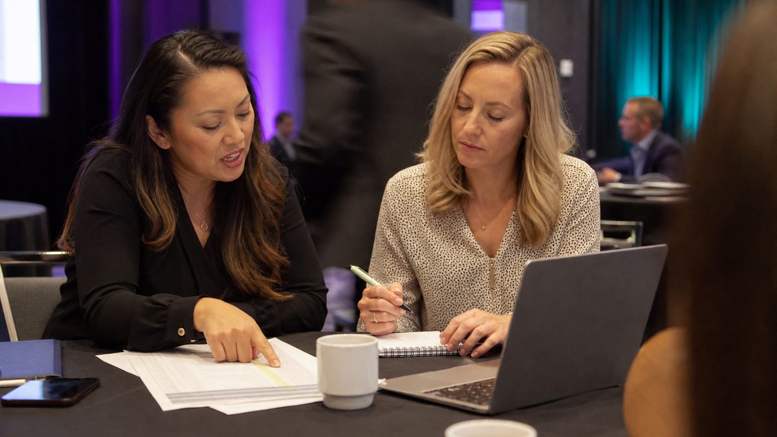 Two corporate event planners across a hotel meeting-room table mid-discussion