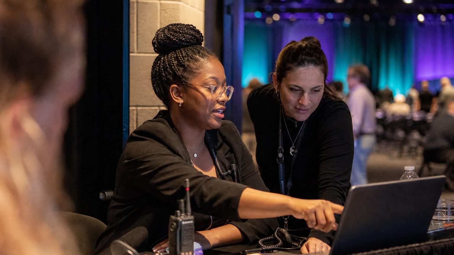 Two event operations colleagues at a laptop in a venue back-of-house area during an event