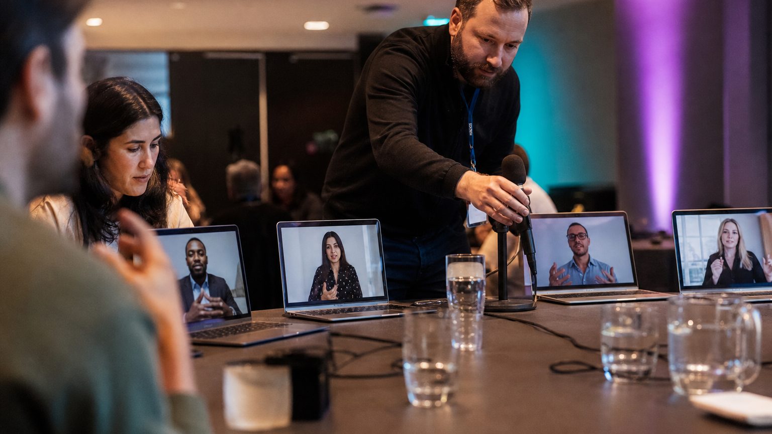 Facilitator adjusts a microphone at a conference table while four remote participants appear on individual laptop scre