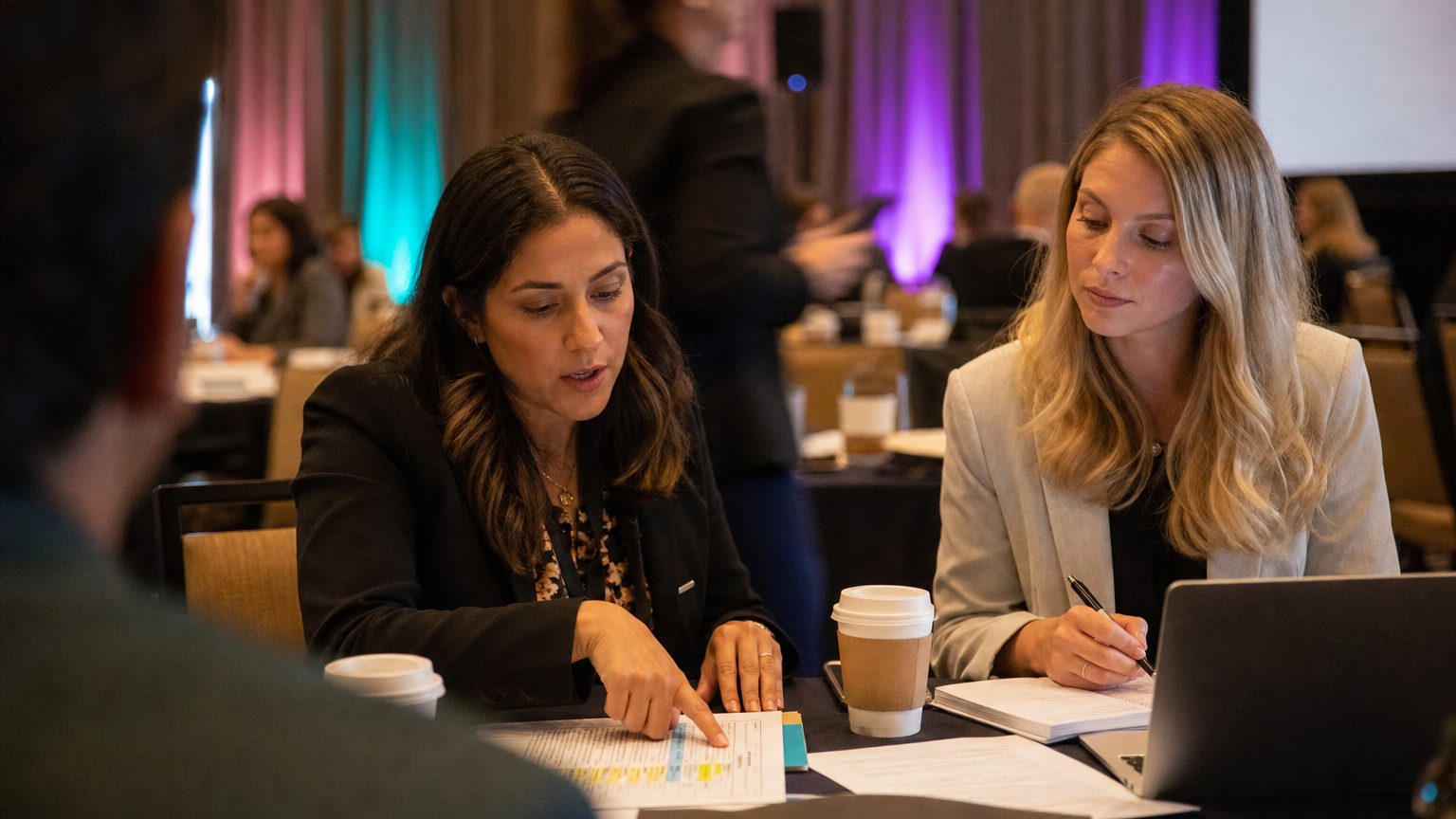 Two corporate event planners across a hotel meeting-room table mid-discussion