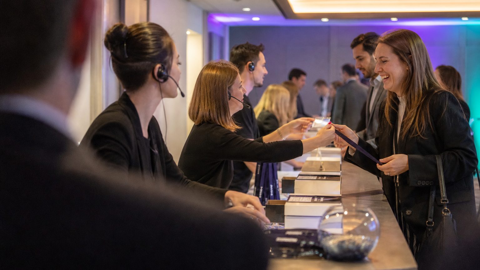Three staff in headsets handing badges across the counter to arriving attendees
