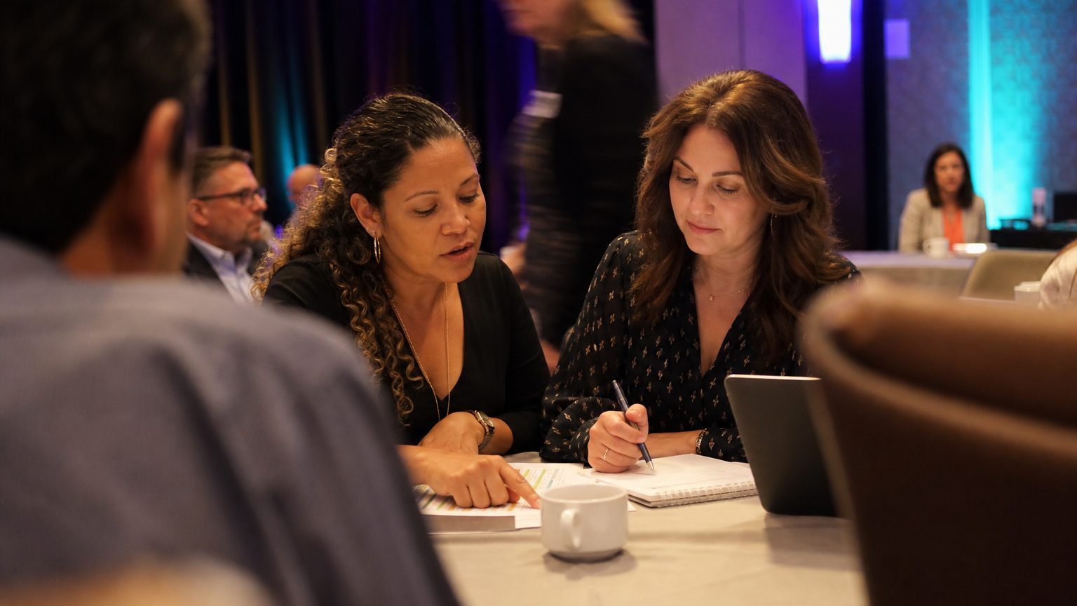 Two corporate event planners across a hotel meeting-room table mid-discussion