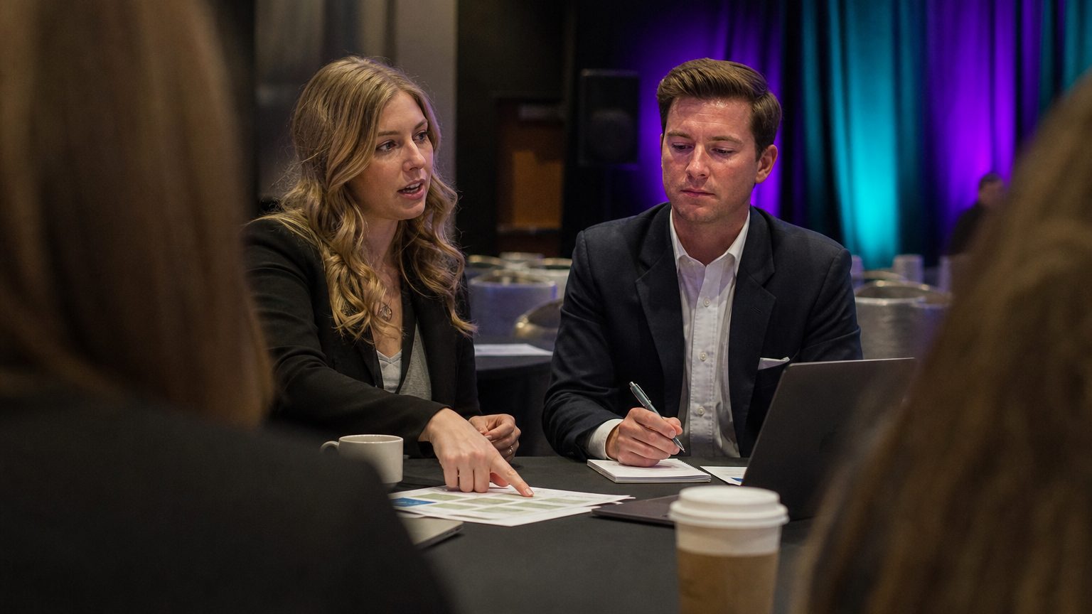Two corporate event planners across a hotel meeting-room table mid-discussion