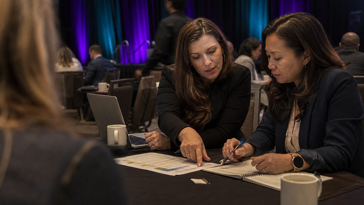 Two corporate event planners across a hotel meeting-room table mid-discussion