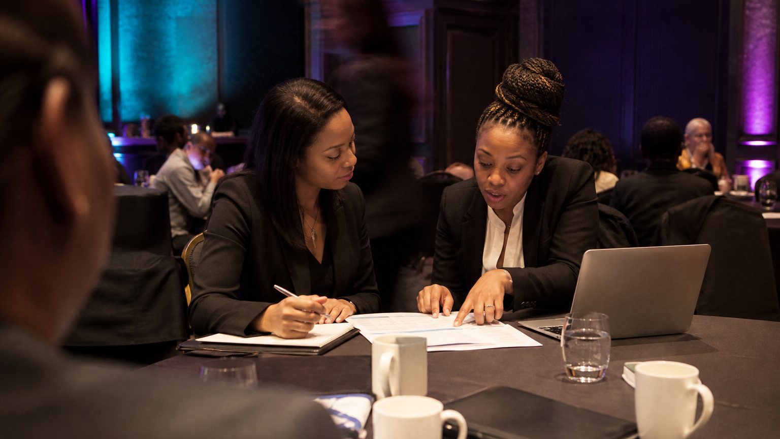 Two corporate event planners across a hotel meeting-room table mid-discussion