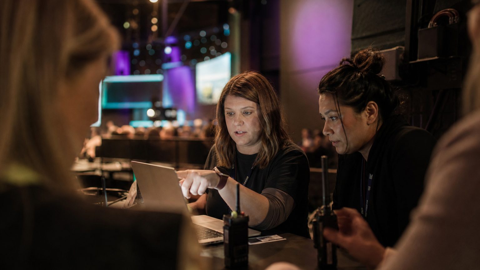 Two event operations colleagues at a laptop in a venue back-of-house area during an event