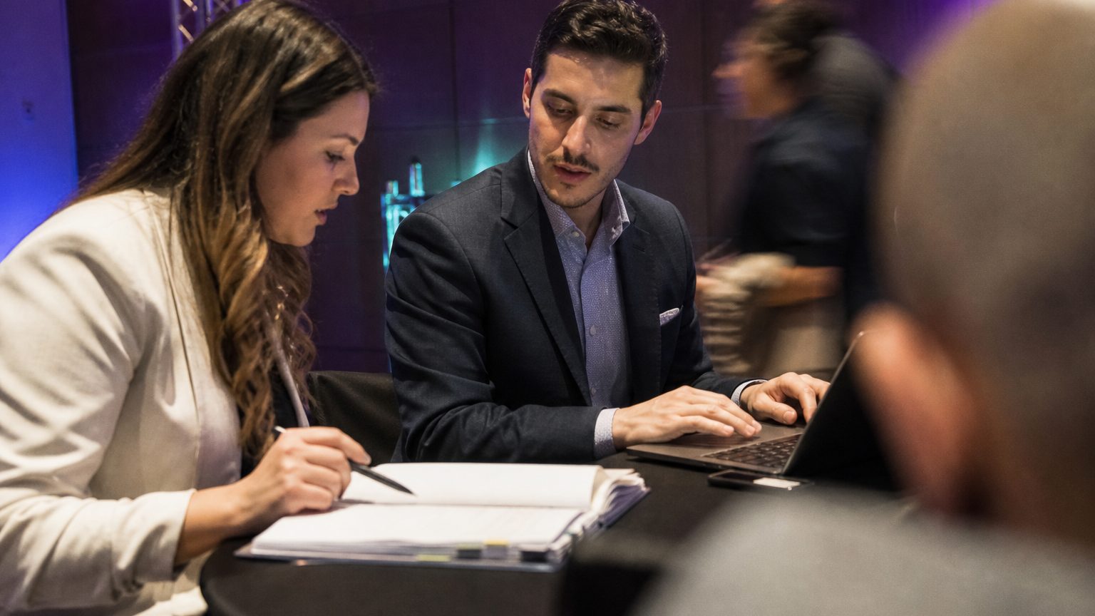 Close-up of two professionals sitting at a table reviewing a stack of event proposals and contract documents, one poin