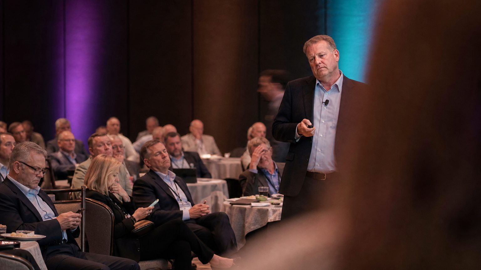 Tired presenter frowns while reading dull slides to a disengaged audience in a dim conference room, some attendees che