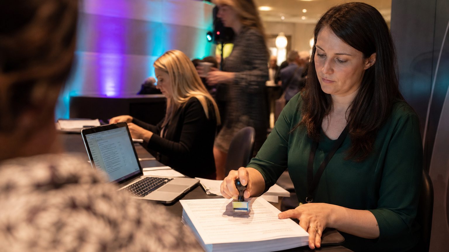 Event coordinator at a desk stamps a stack of attendee data forms labeled “Confidential,” while a laptop screen shows 