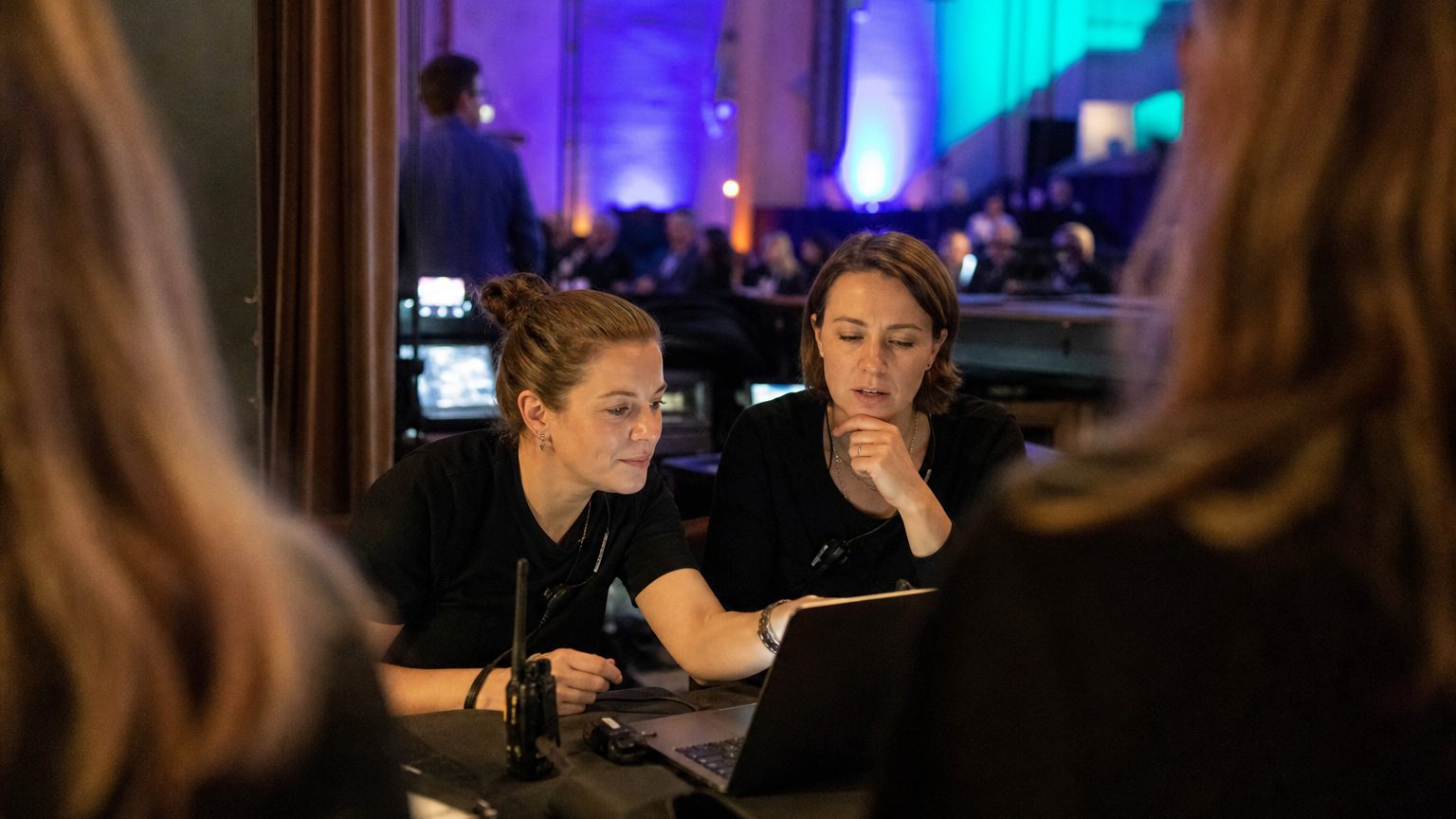 Two event operations colleagues at a laptop in a venue back-of-house area during an event