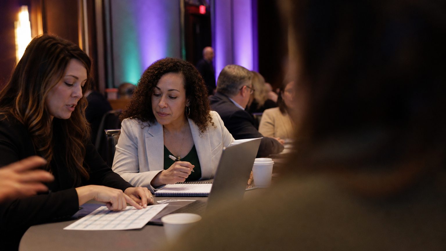 Two corporate event planners across a hotel meeting-room table mid-discussion