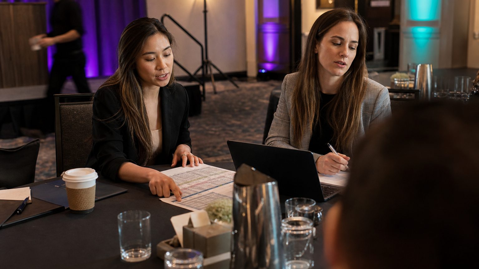 Two corporate event planners across a hotel meeting-room table mid-discussion