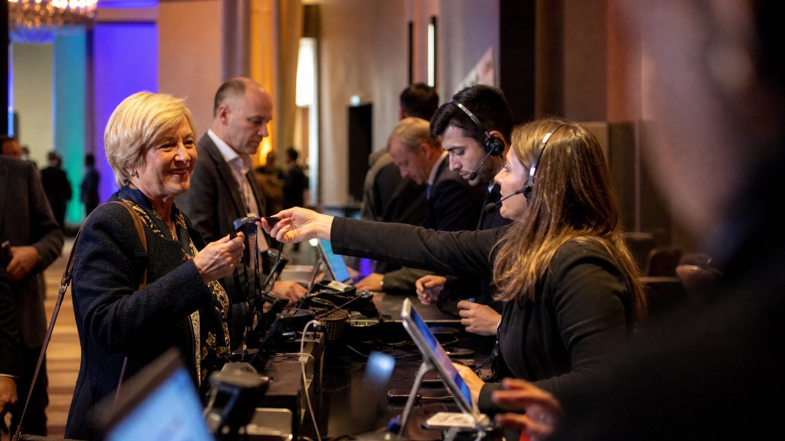 Three staff in headsets handing badges across the counter to arriving attendees