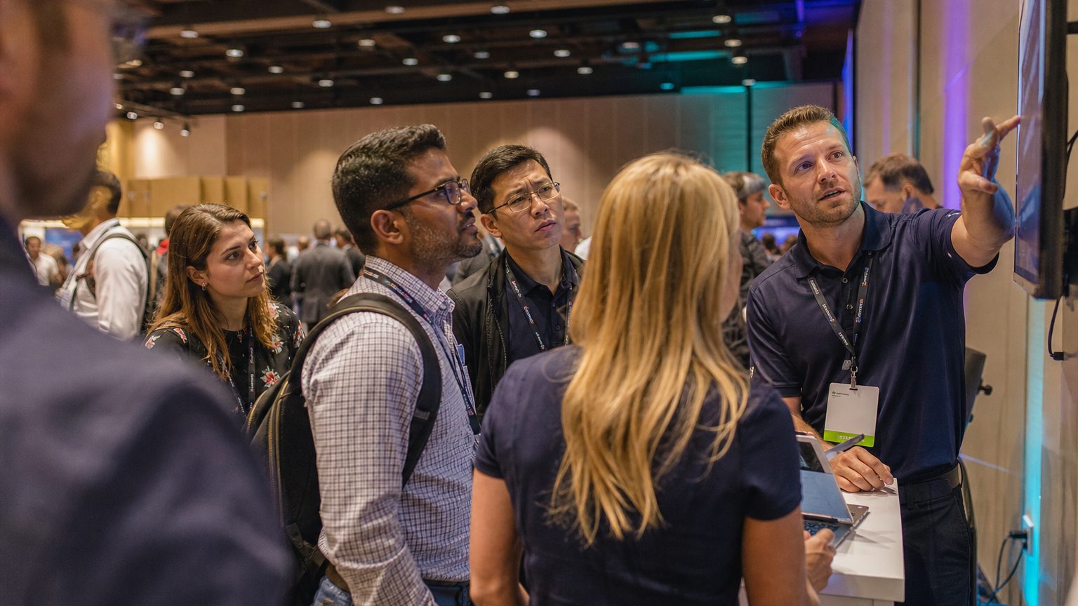 Two exhibitors in branded polos talking with three visitors at a booth counter