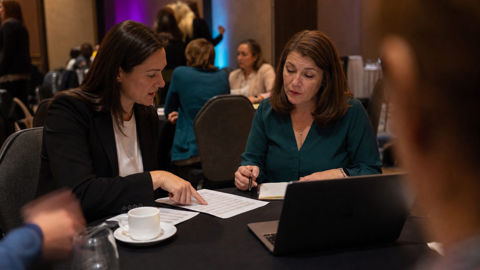 Two corporate event planners across a hotel meeting-room table mid-discussion