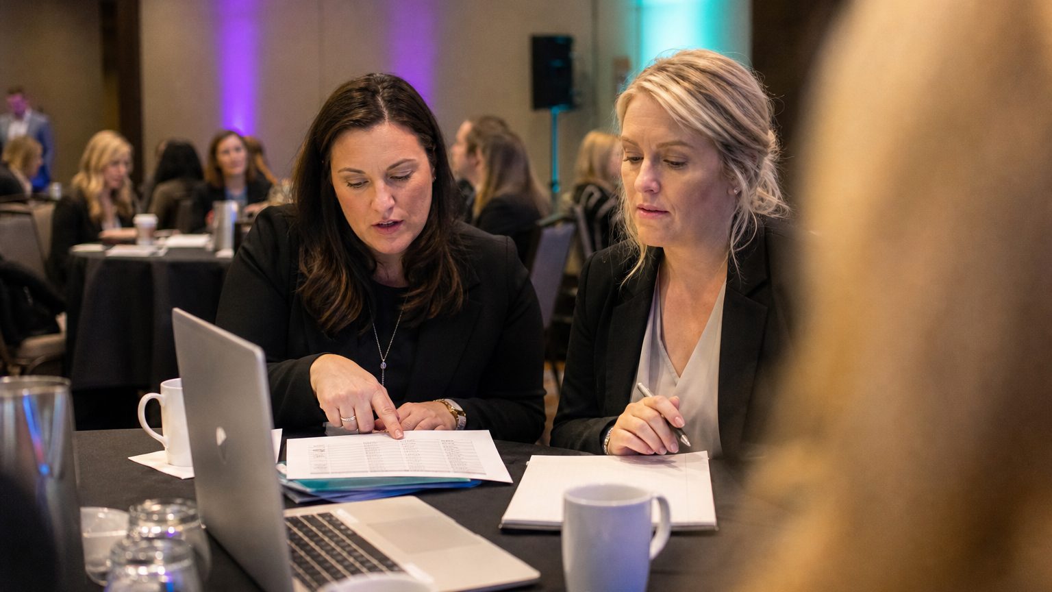 Two corporate event planners across a hotel meeting-room table mid-discussion