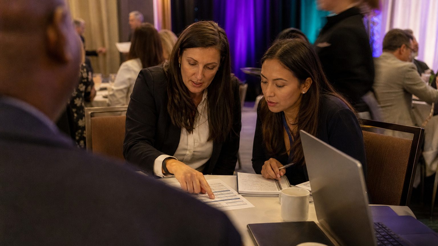 Two corporate event planners across a hotel meeting-room table mid-discussion