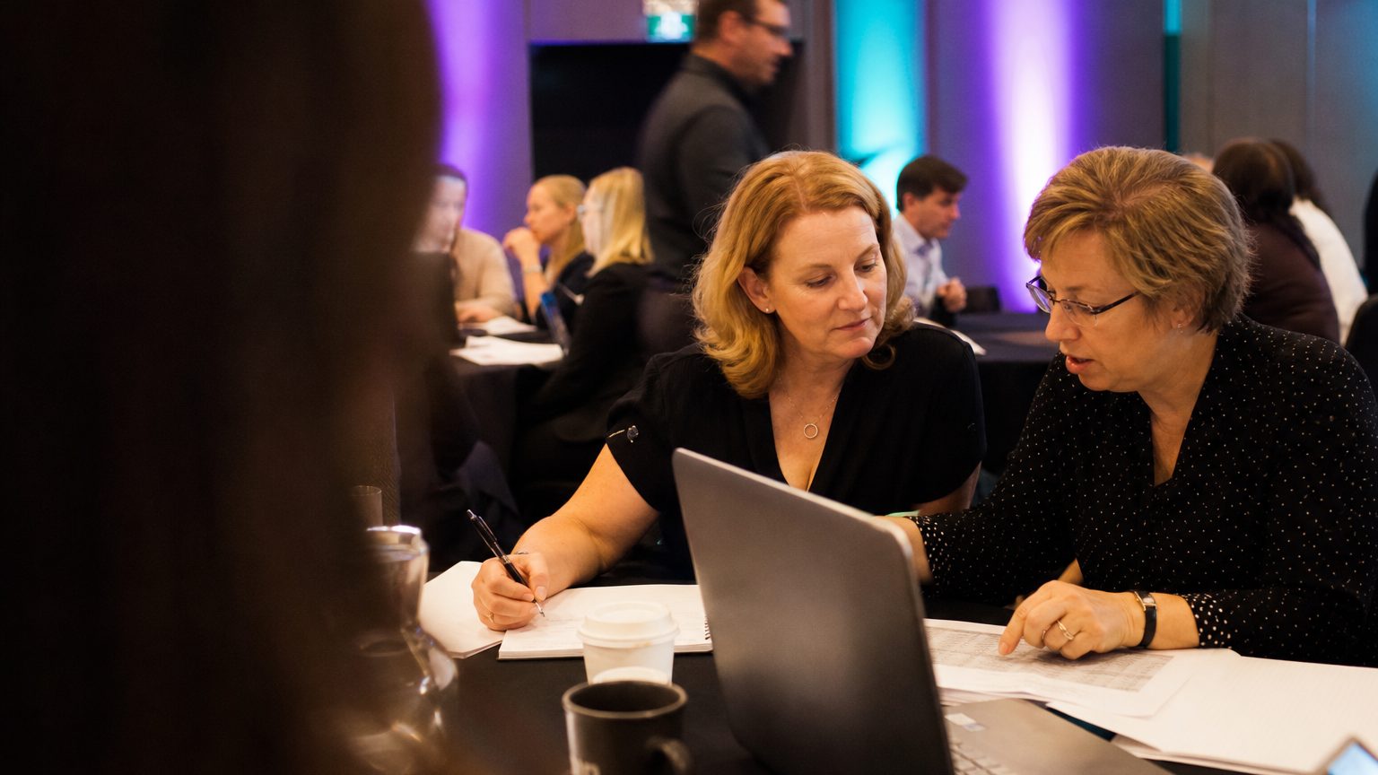 Two corporate event planners across a hotel meeting-room table mid-discussion