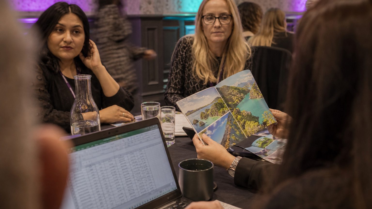 Close-up of a planner’s hands flipping through a colorful brochure featuring exotic destinations, with a laptop showin