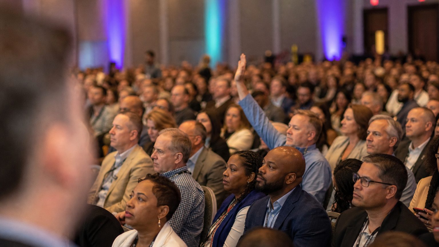 A presenter gesturing at the front of a packed ballroom