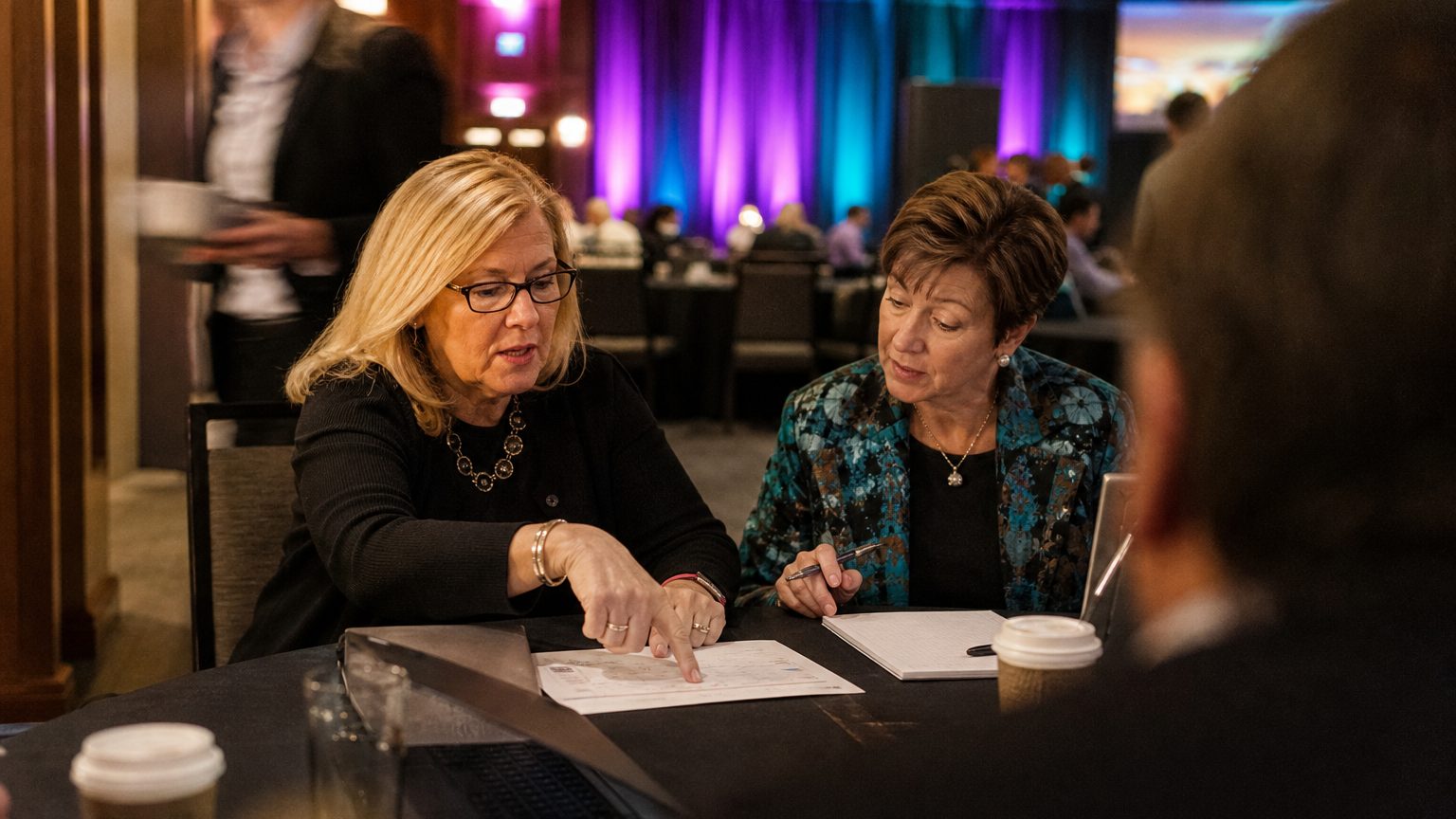 Two corporate event planners across a hotel meeting-room table mid-discussion