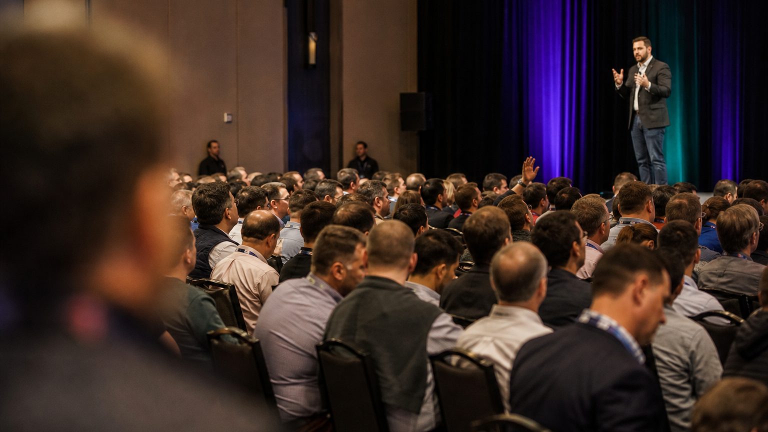 A presenter gesturing at the front of a packed ballroom