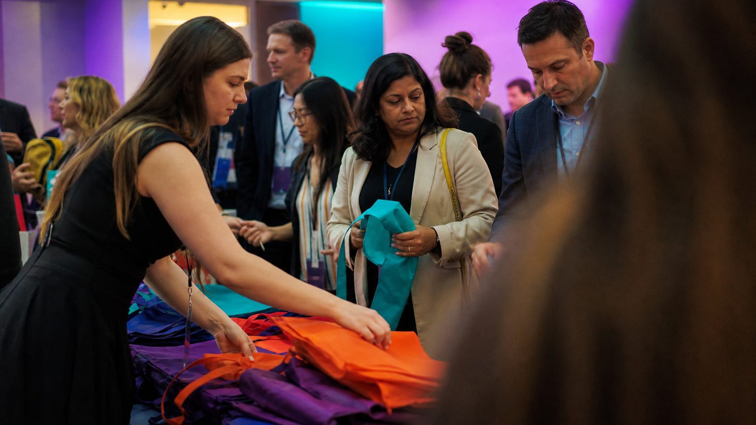 Close-up of hands arranging brightly colored branded tote bags and lanyards on a table, with attendees reaching to pic
