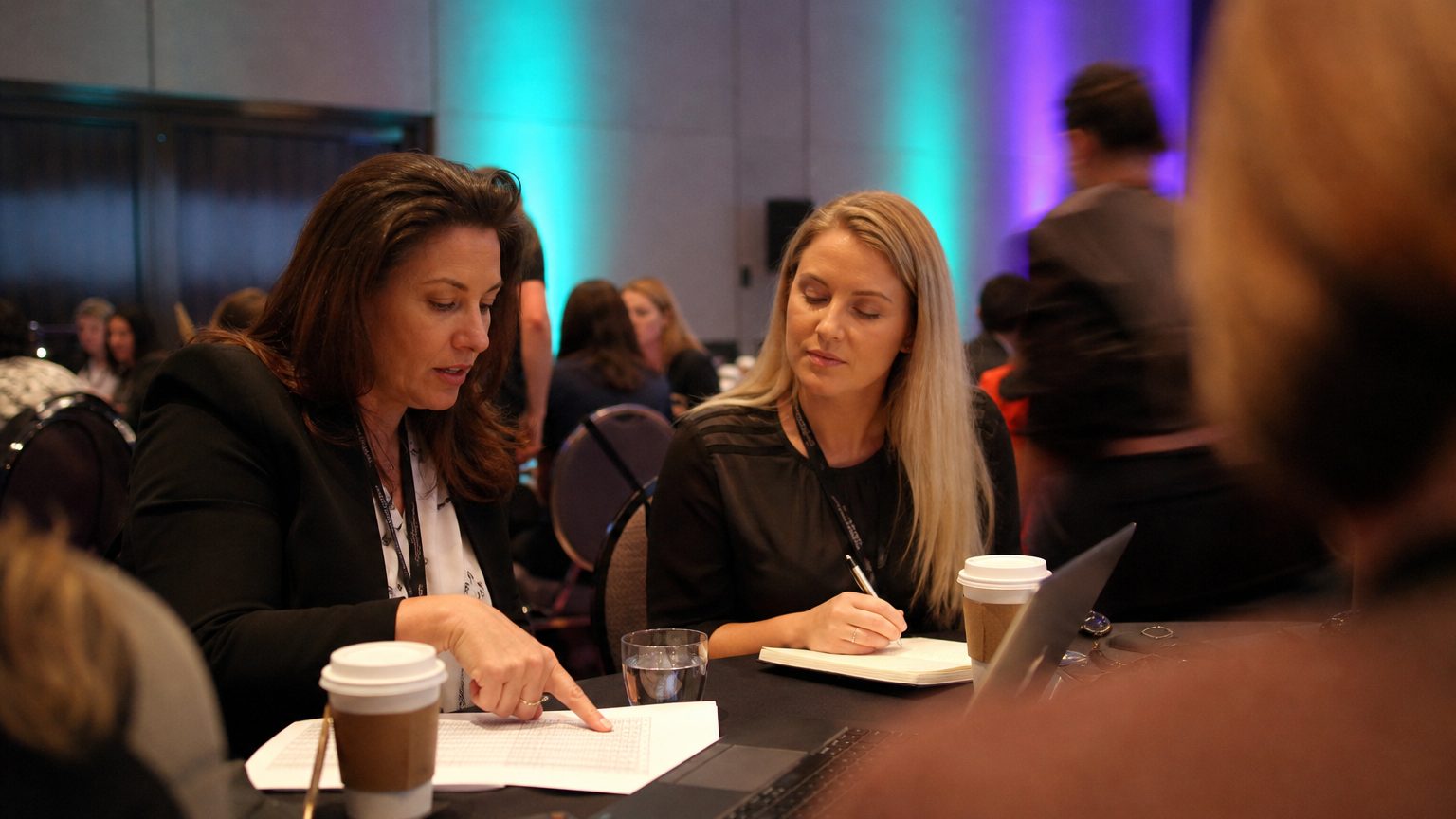 Two corporate event planners across a hotel meeting-room table mid-discussion