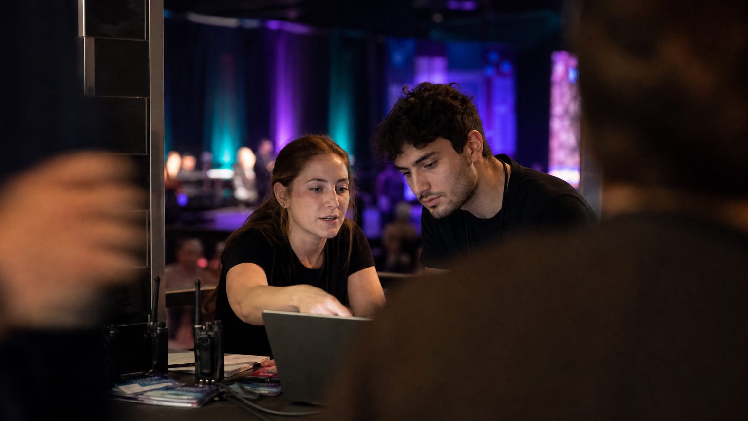 Two event operations colleagues at a laptop in a venue back-of-house area during an event