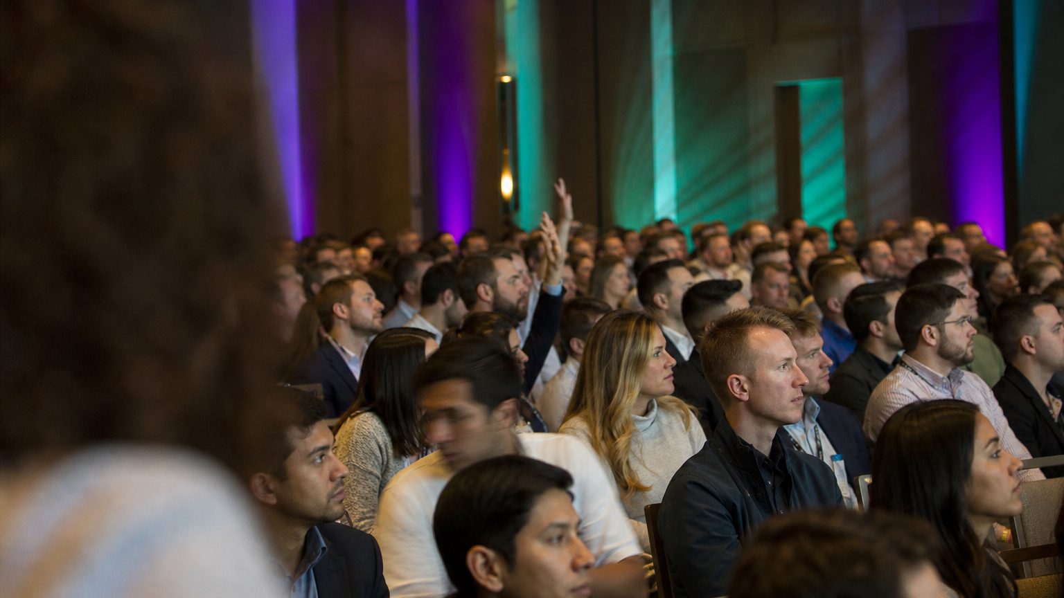 A presenter gesturing at the front of a packed ballroom