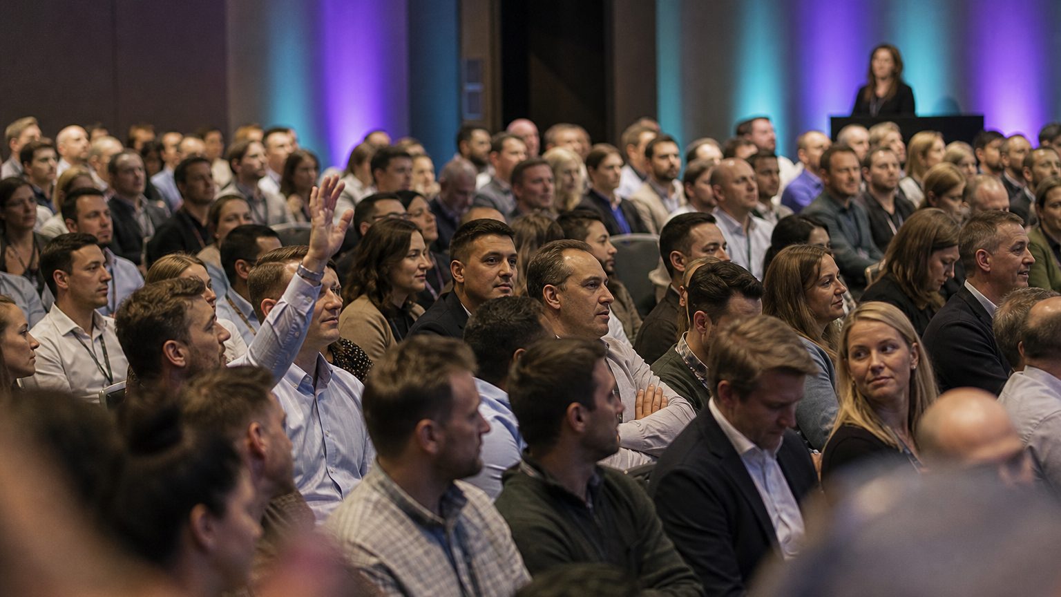 A presenter gesturing at the front of a packed ballroom