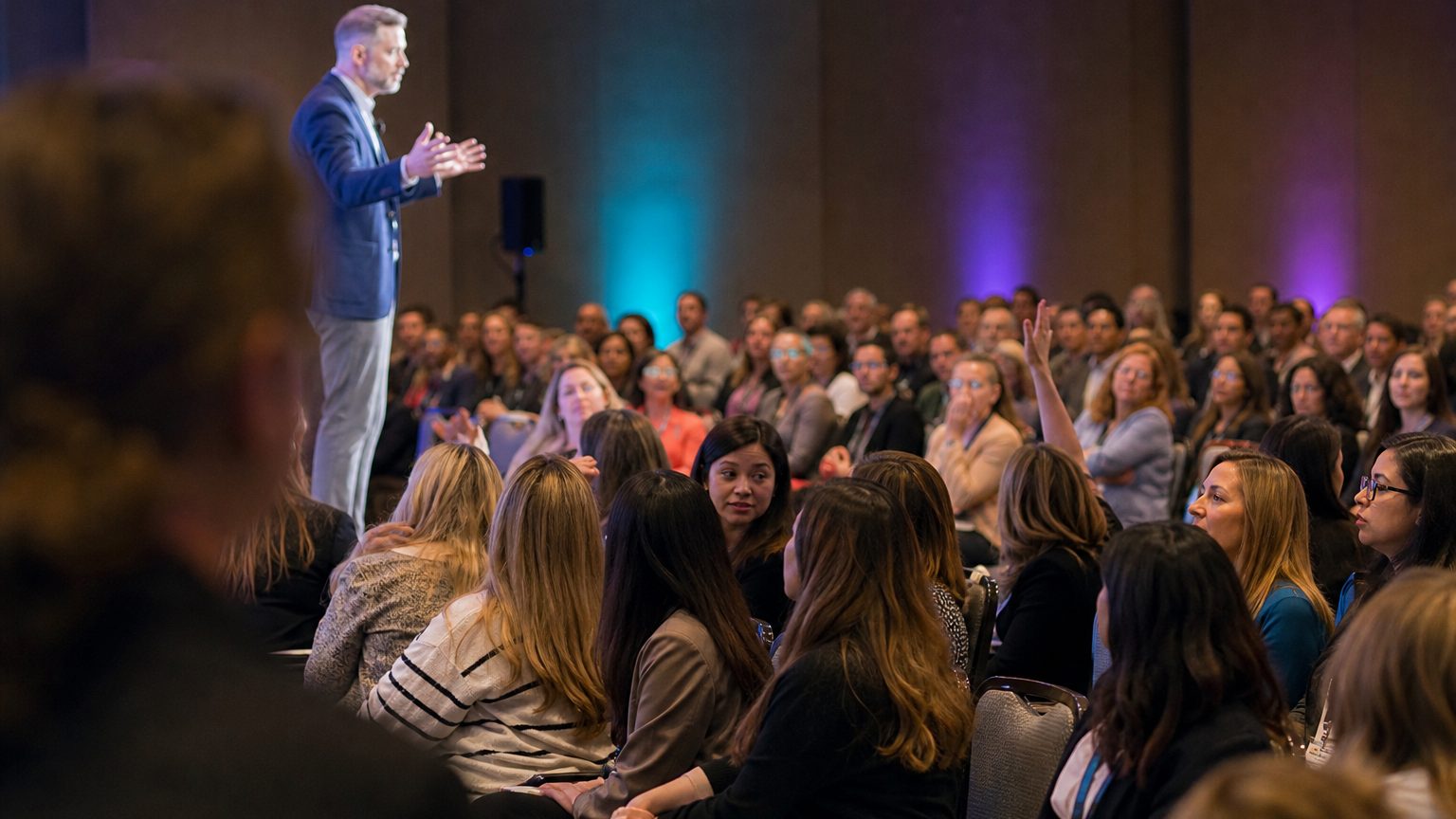 A presenter gesturing at the front of a packed ballroom