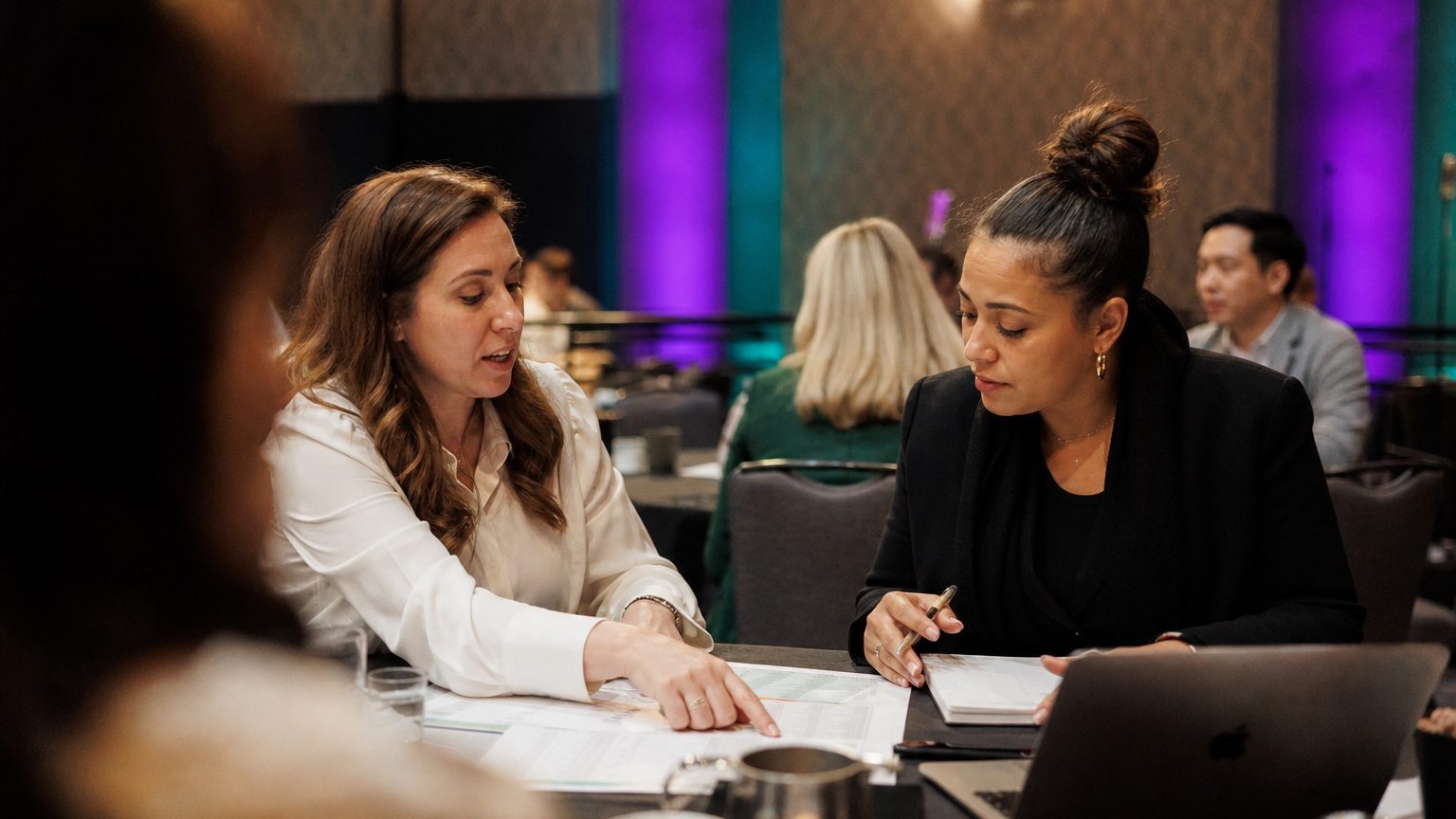 Two corporate event planners across a hotel meeting-room table mid-discussion
