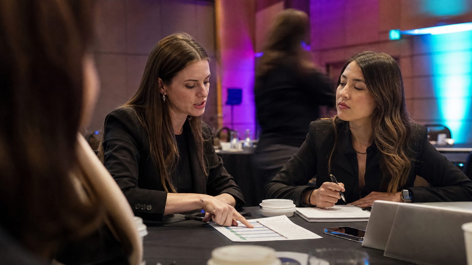 Two corporate event planners across a hotel meeting-room table mid-discussion