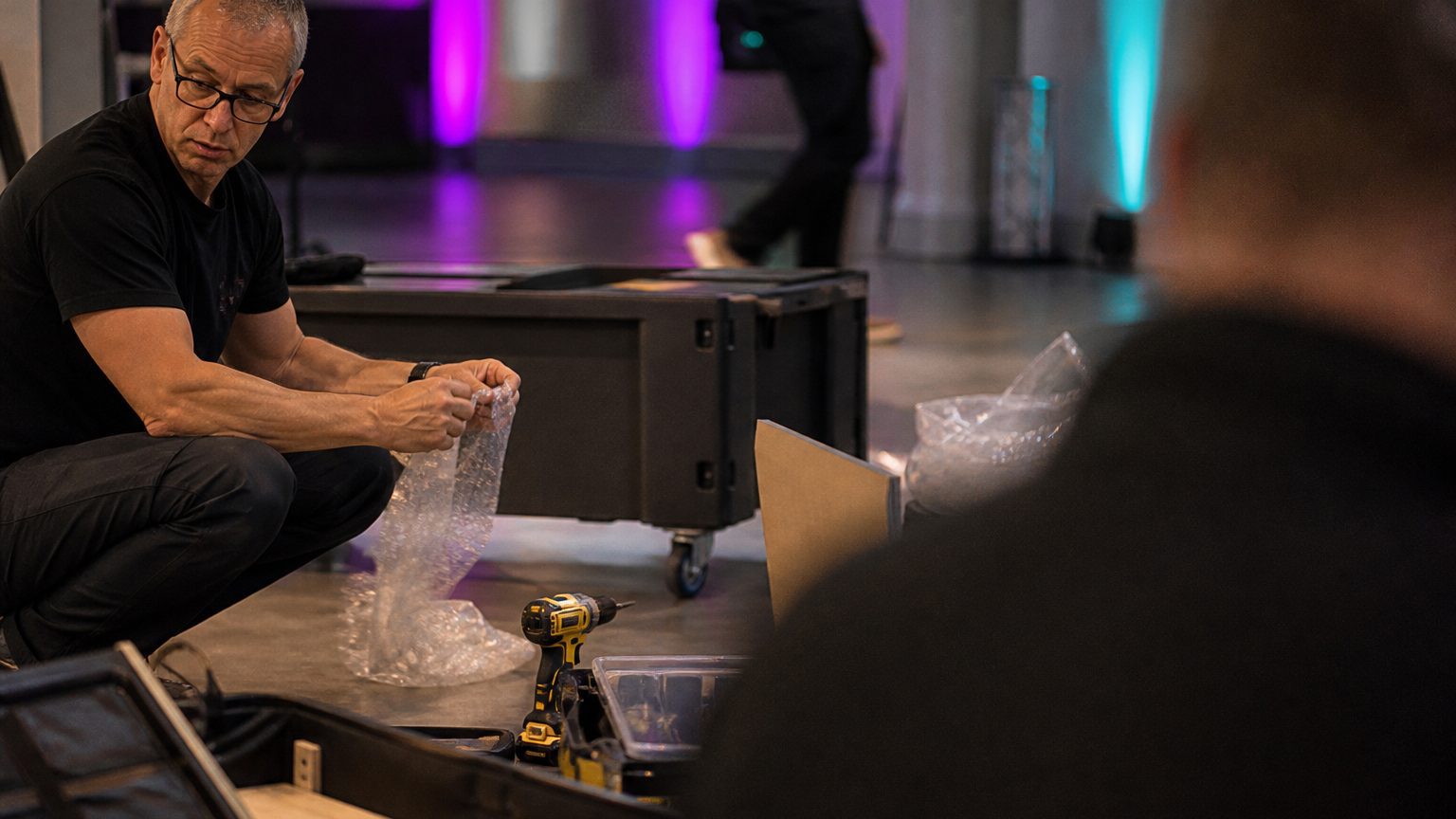 Close-up of a trade show worker unpacking booth crates on a concrete floor, with tools and packaging materials scatter