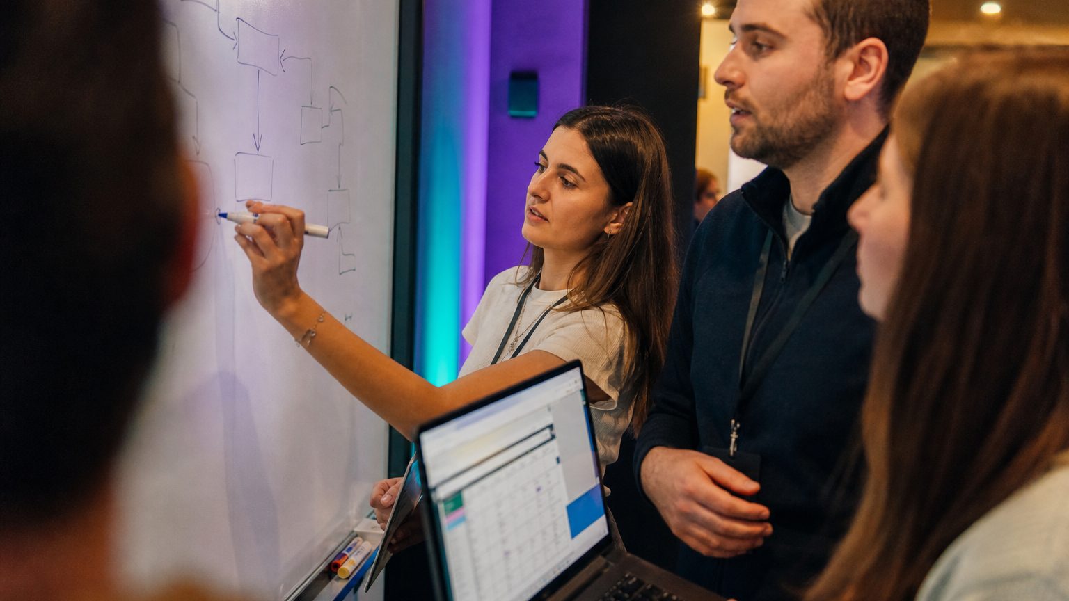 Close-up of hands writing notes and drawing a flowchart on a whiteboard, with a laptop open nearby displaying a simple