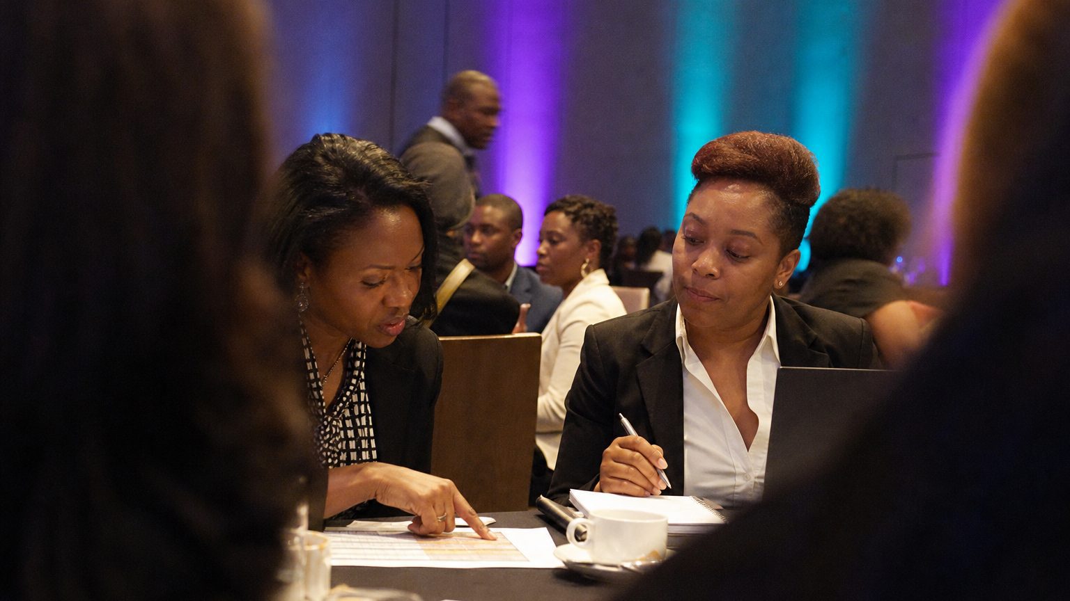Two corporate event planners across a hotel meeting-room table mid-discussion