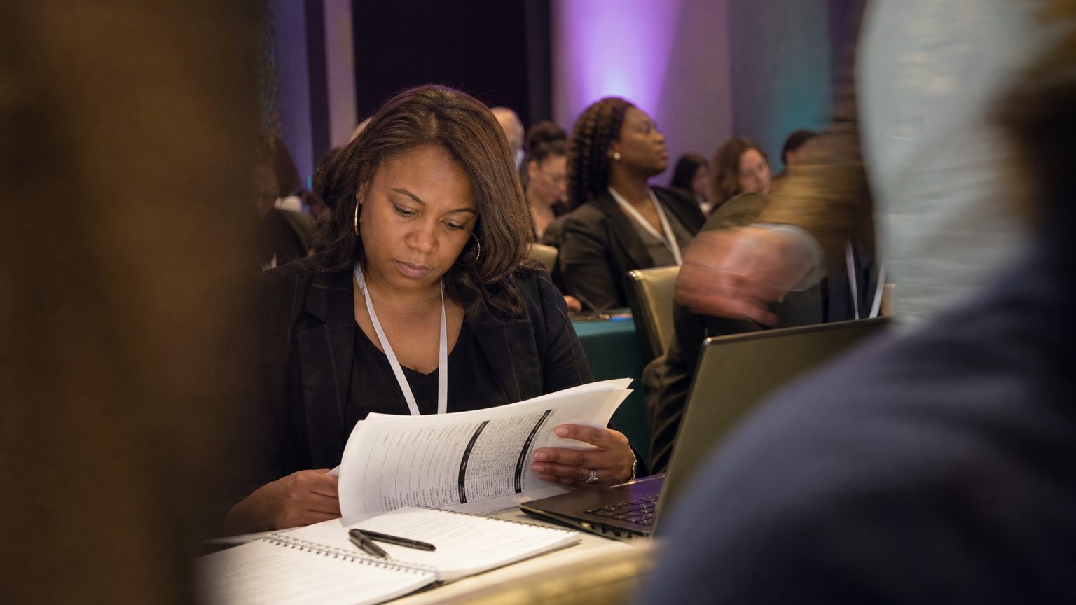 Close-up of a professional reviewing a printed certification exam booklet at a desk cluttered with notes and a laptop,