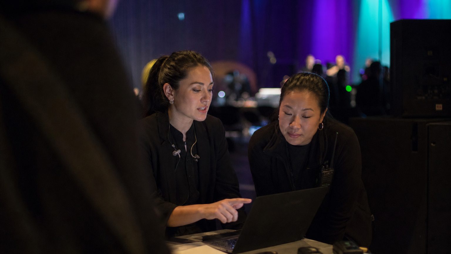 Two crew members at the front-of-house position with stage lighting behind them