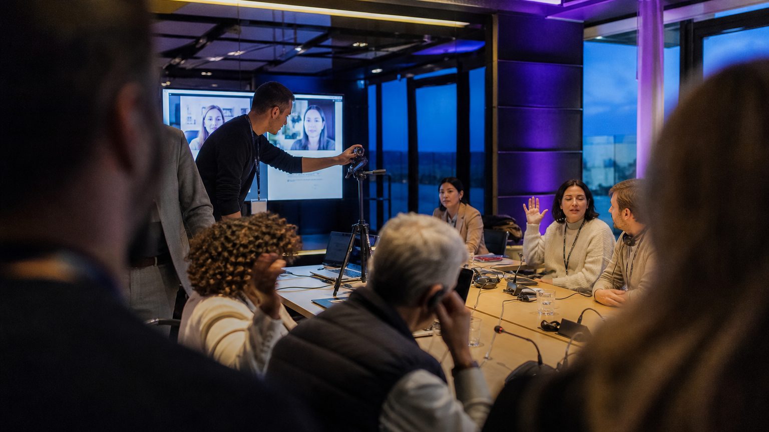 Facilitator adjusts a laptop camera on a tripod as remote and in-person participants interact during a hybrid team-bui