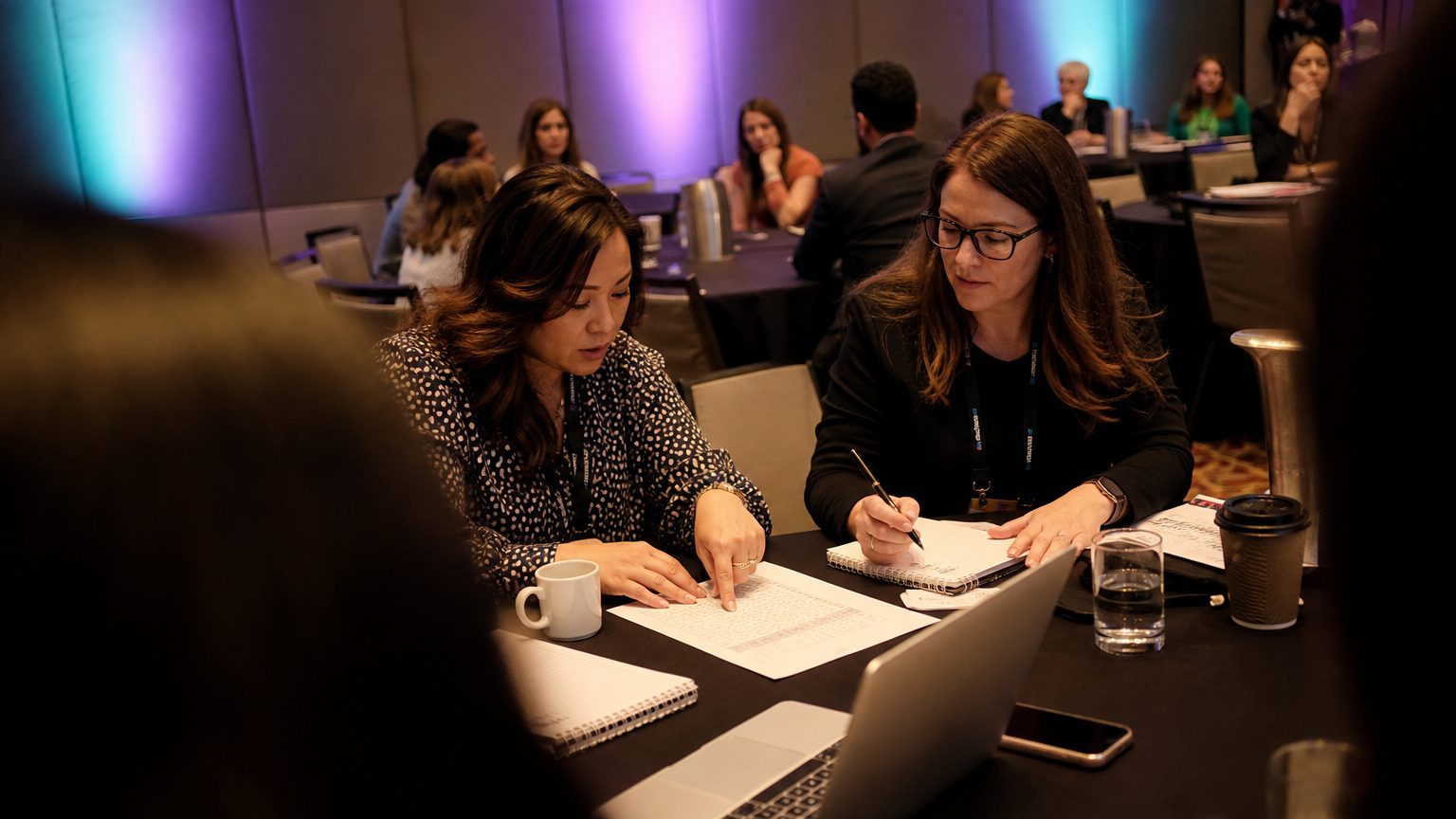Two corporate event planners across a hotel meeting-room table mid-discussion