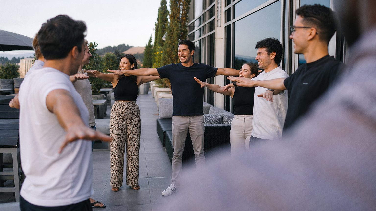 A group of eight people stand in a loose circle on a hotel terrace in afternoon light, mid-laugh, one participant holding both arms out horizontally as the others mirror the pose with varying degrees of commitment.
