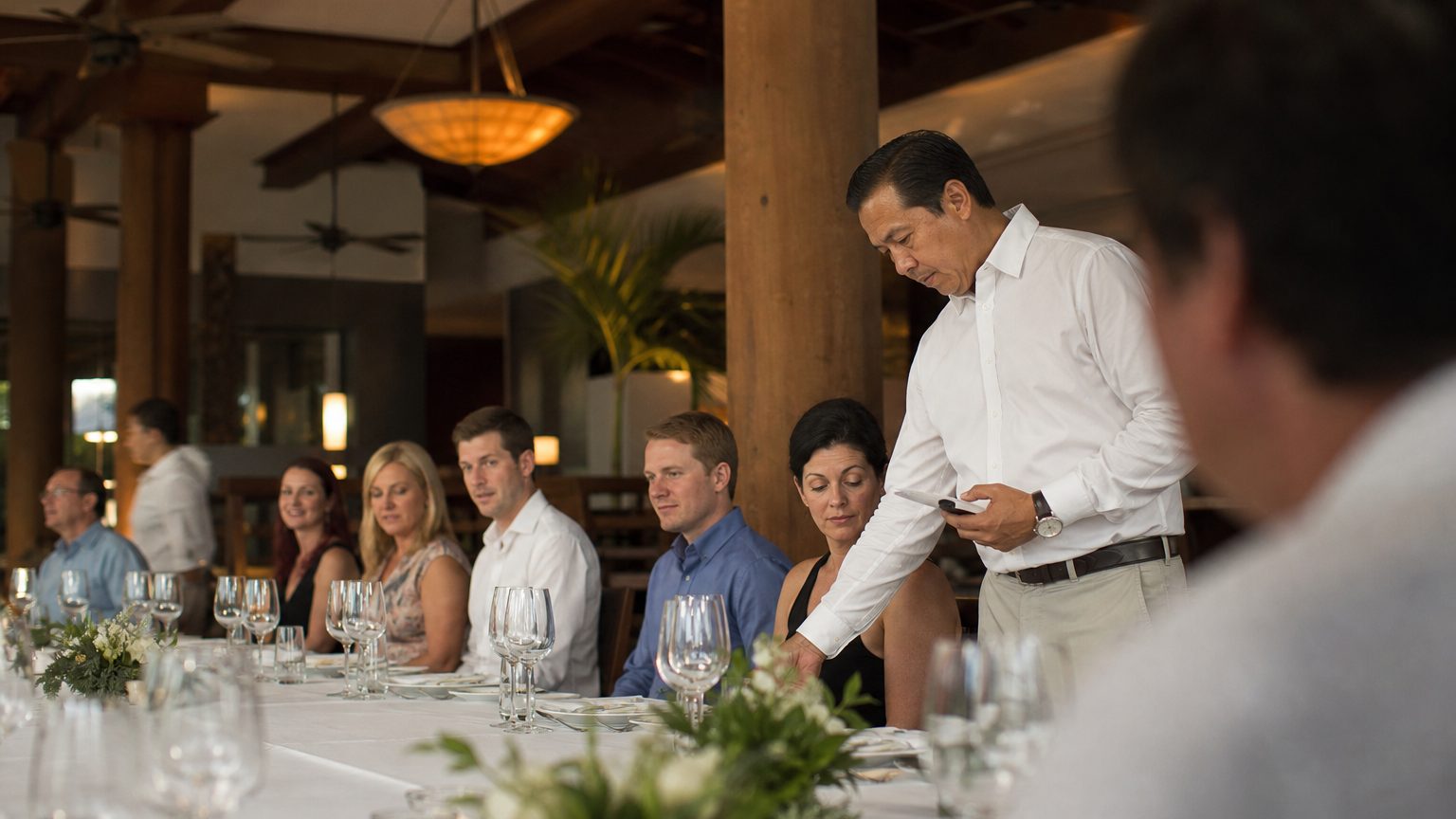 A catering manager in a resort dining room counts place settings along a long table laid for a formal dinner, late-afternoon light falling across white linen and glassware.