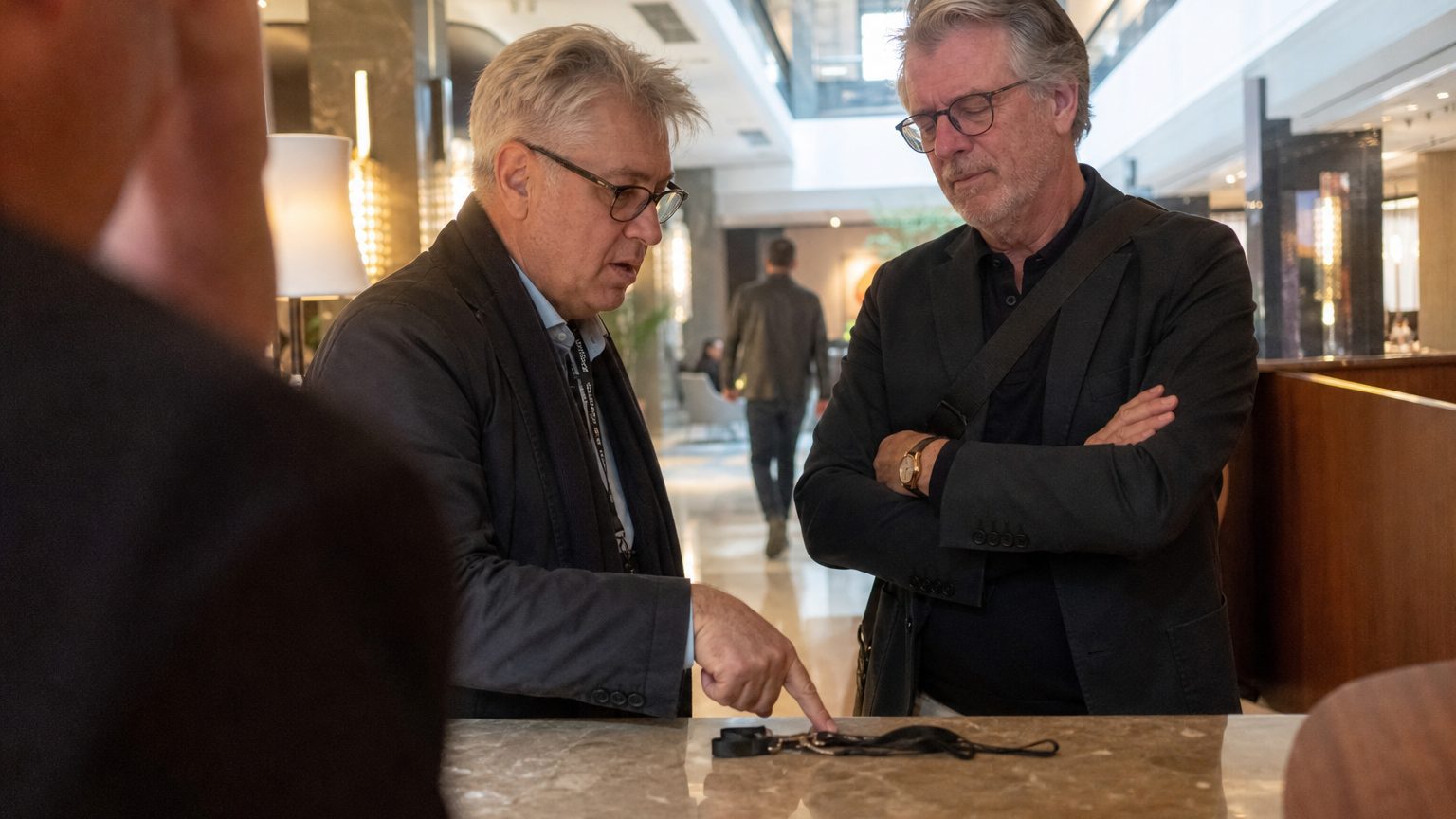 A tax adviser and an event producer stand at a hotel concierge desk in a marble-floored lobby, the adviser pointing at a lanyard on the counter while explaining something, the producer listening with arms crossed, midday light from a skylight overhead.