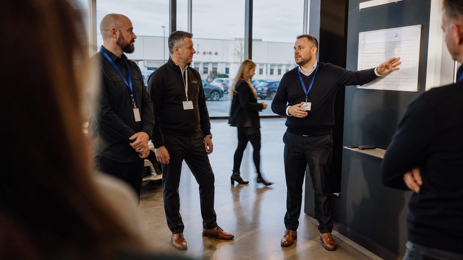 A sales manager in an open-plan showroom floor stands at a branded product display, gesturing toward a mounted quota tracker board while two colleagues in lanyards listen, afternoon light through floor-to-ceiling windows casting long shadows across the polished concrete.