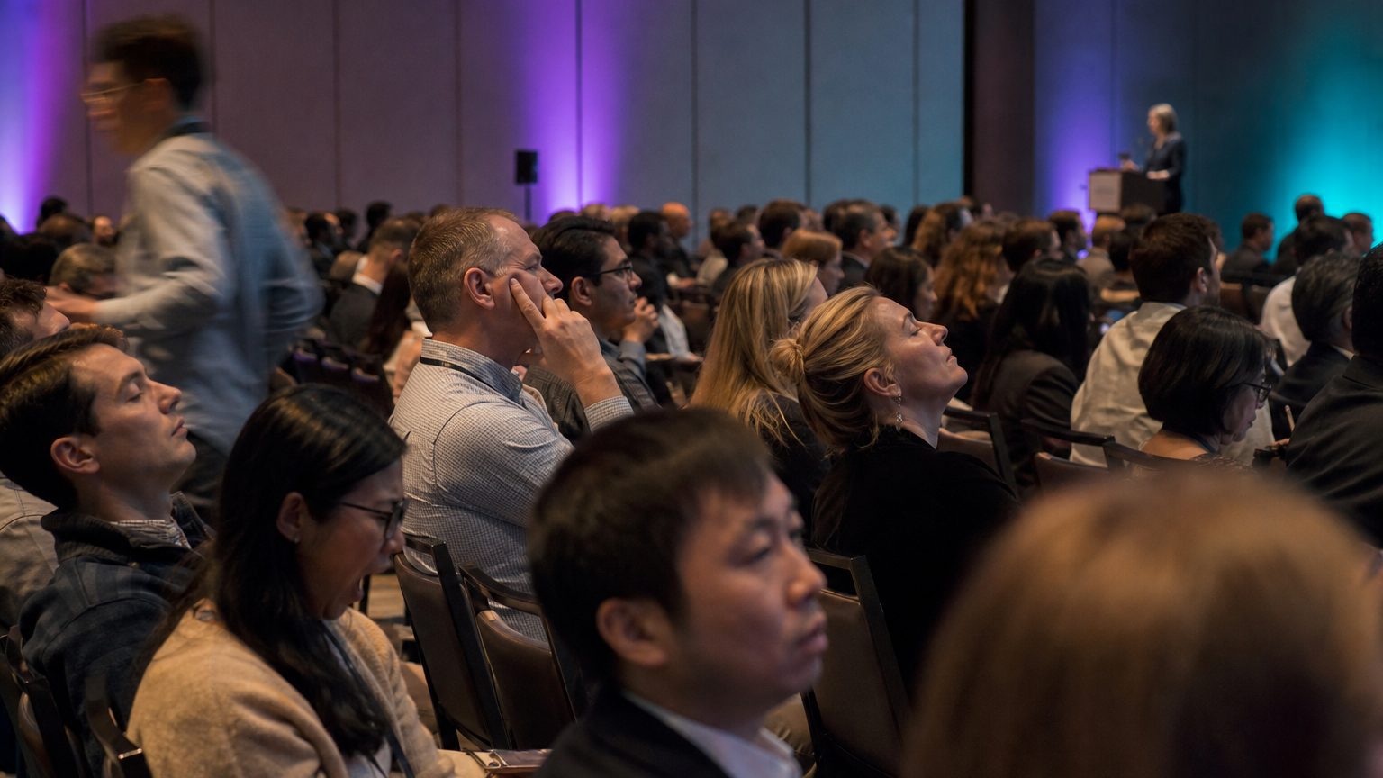 A wide conference room filled with rows of seated employees, several visibly slumped in chairs or staring at the ceiling while a presenter speaks at a distant podium under flat overhead lighting.