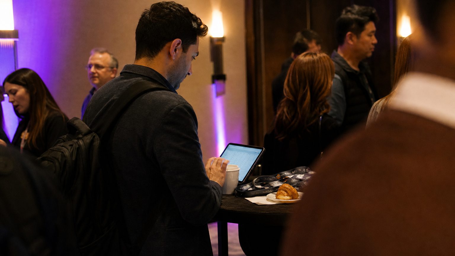 A media buyer stands at a tall bar table in a hotel corridor outside a closed session room, tapping through a campaign dashboard on a tablet propped against a coffee cup, a stack of conference lanyards and a half-eaten pastry beside them.