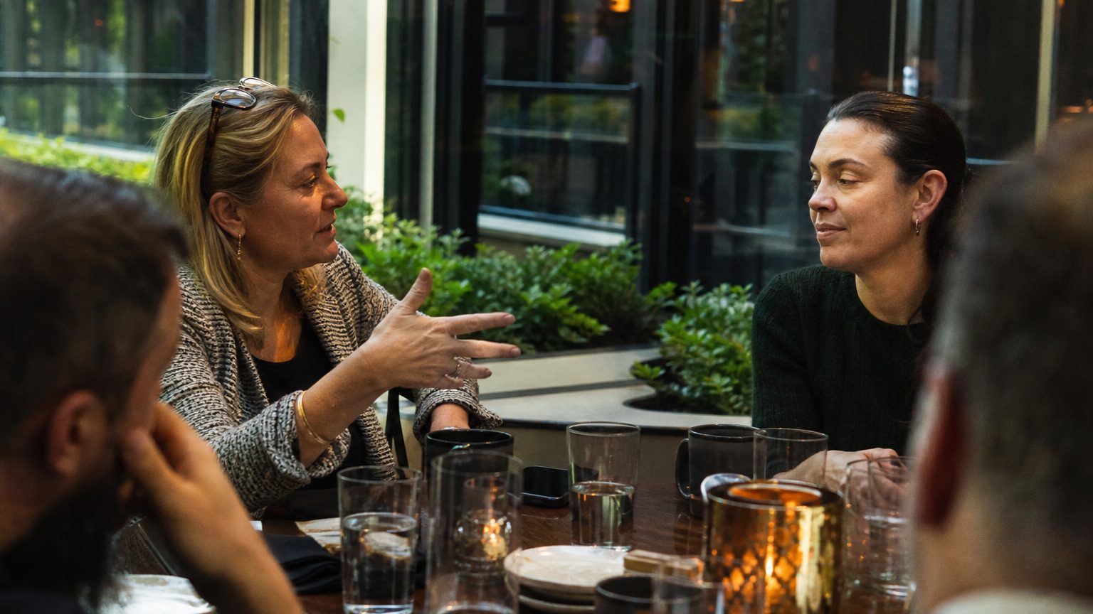 A payroll manager and a travel program director sit across from each other at a restaurant table, one gesturing toward the other mid-conversation, coffee cups and a smartphone between them, late morning light coming through a window beside them.