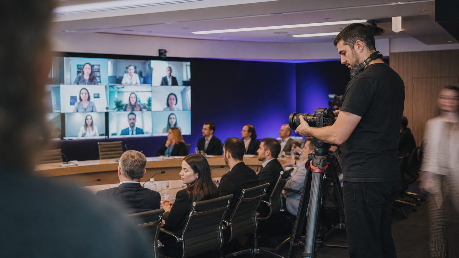 A conference room camera operator adjusts a wide-angle lens on a tripod aimed at a horseshoe-shaped table where in-person attendees sit facing a wall of mounted screens showing remote participants in individual video tiles.