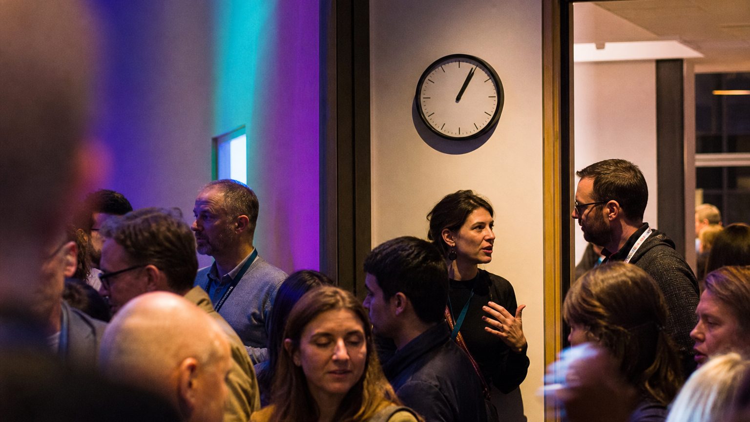 A facilitator stands at the edge of a breakout room doorway, lanyard over one shoulder, speaking directly to a colleague in the corridor while a wall clock above them shows the time just past the hour.